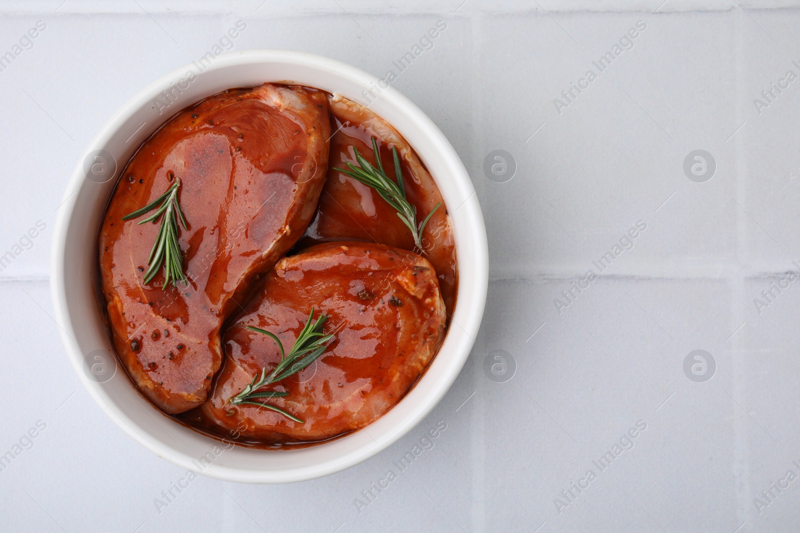 Photo of Raw marinated meat and rosemary in bowl on white tiled table, top view. Space for text