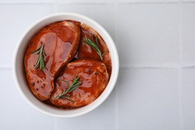 Raw marinated meat and rosemary in bowl on white tiled table, top view. Space for text