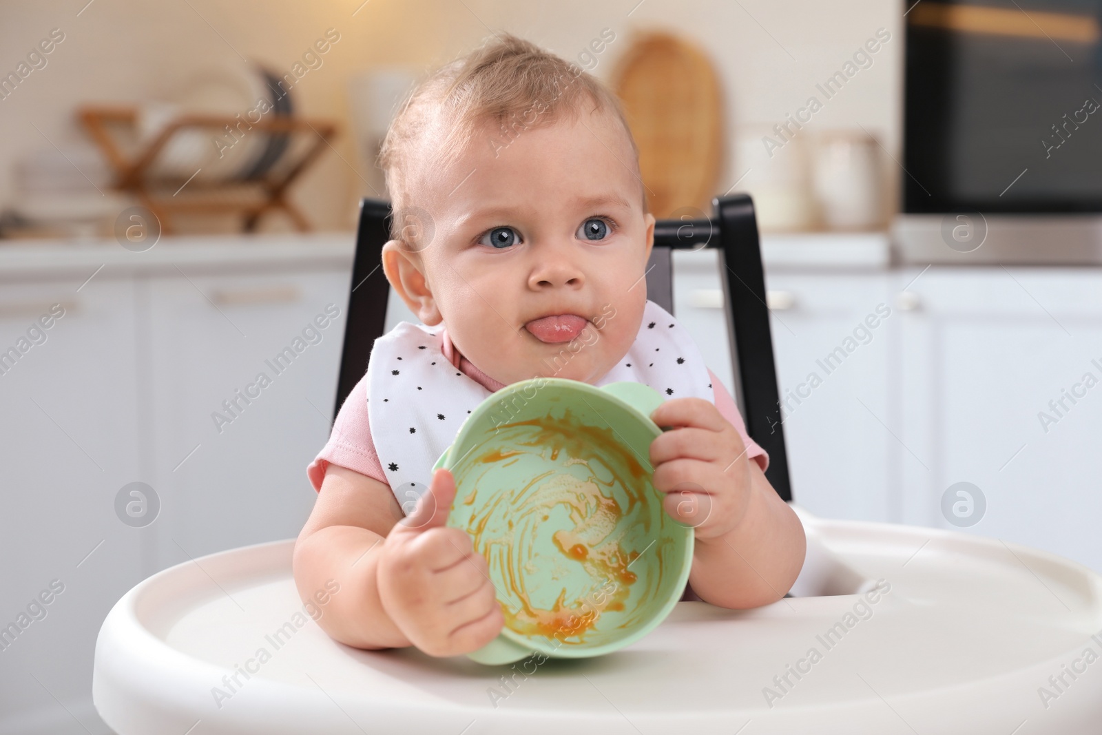 Photo of Cute little baby with bowl in high chair at kitchen