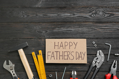 Photo of Card with phrase HAPPY FATHER'S DAY and different tools on black wooden background, flat lay. Space for text