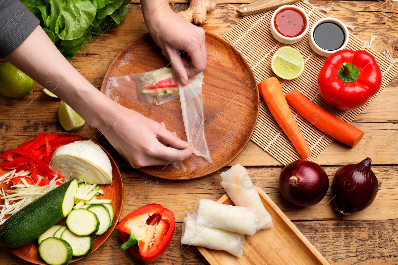 Photo of Making delicious spring rolls. Woman wrapping fresh vegetables into rice paper at wooden table, flat lay