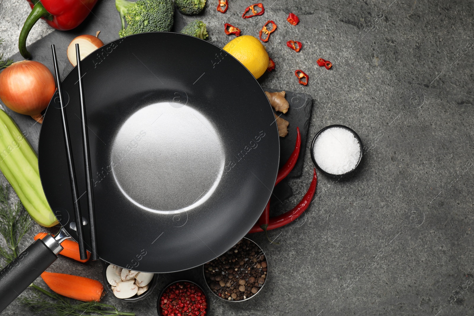 Photo of Empty iron wok and chopsticks surrounded by ingredients on grey table, flat lay. Space for text