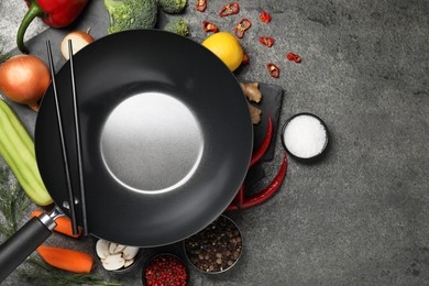 Empty iron wok and chopsticks surrounded by ingredients on grey table, flat lay. Space for text