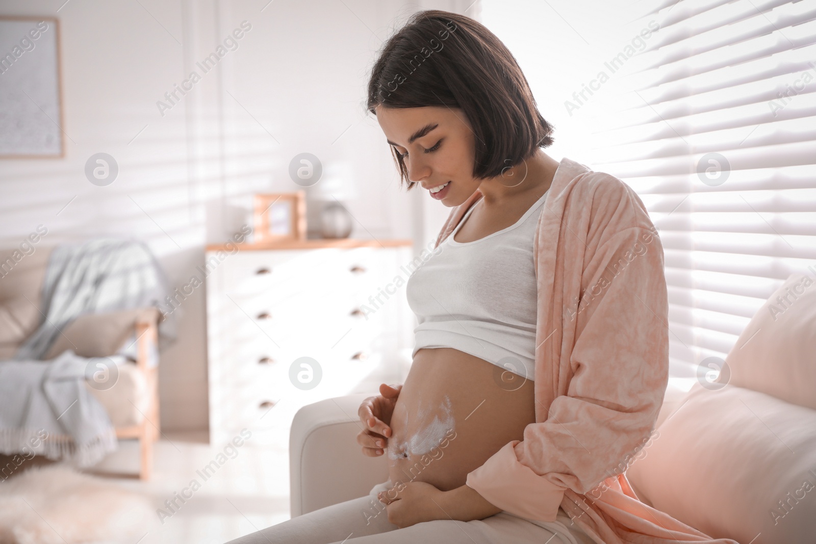 Photo of Young pregnant woman applying cosmetic product on belly at home