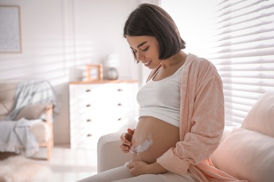 Photo of Young pregnant woman applying cosmetic product on belly at home