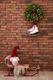 Photo of Christmas gnome on sleigh near brick wall with pair of ice skates and festive wreath indoors