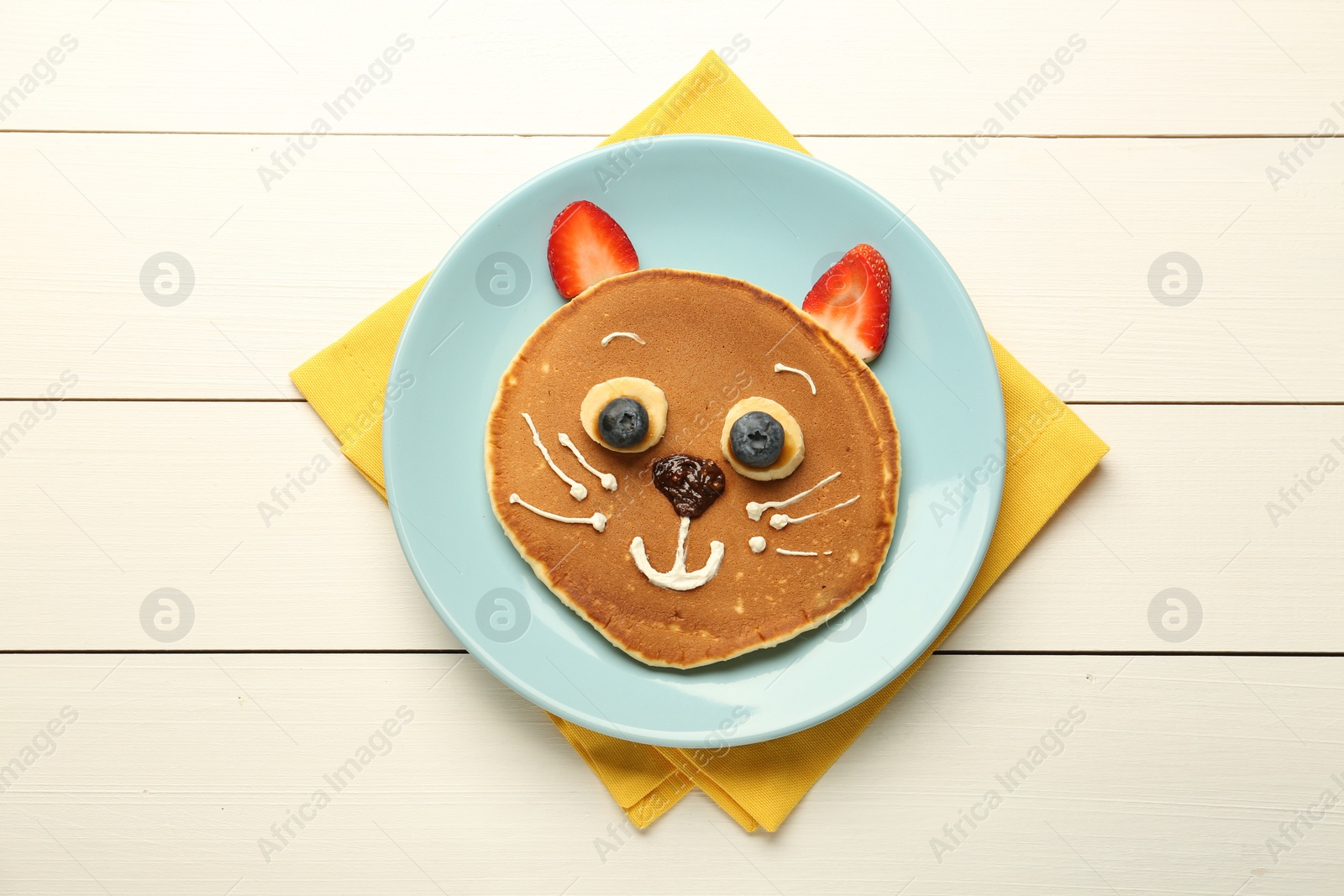 Photo of Creative serving for kids. Plate with cute cat made of pancakes, berries, cream, banana and chocolate paste on white wooden table, top view