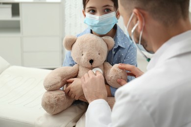 Photo of Pediatrician playing with little girl during visit in hospital. Doctor and patient wearing protective masks