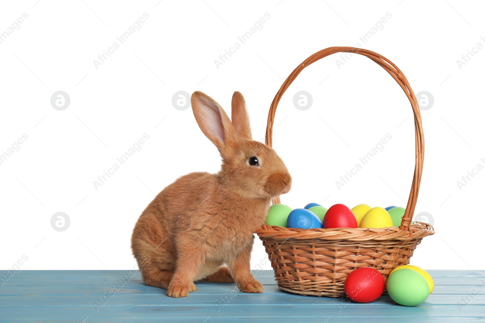 Photo of Cute fluffy bunny and wicker basket with Easter eggs on light blue wooden table