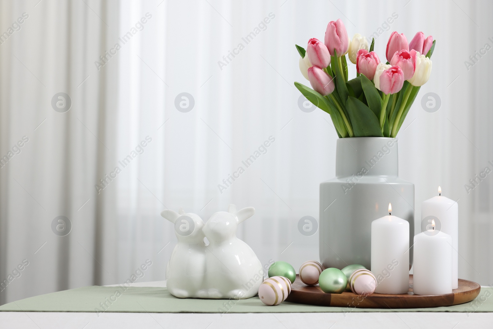 Photo of Easter decorations. Bouquet of tulips, painted eggs, burning candles and bunny figures on white table indoors. Space for text