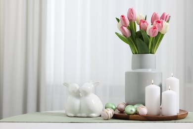 Photo of Easter decorations. Bouquet of tulips, painted eggs, burning candles and bunny figures on white table indoors. Space for text