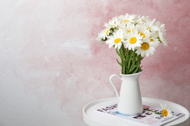 Photo of Jug with beautiful chamomile flowers on table against color background