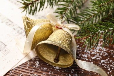 Photo of Golden shiny bells with bow, music sheet and fir branches on wooden table, closeup. Christmas decoration
