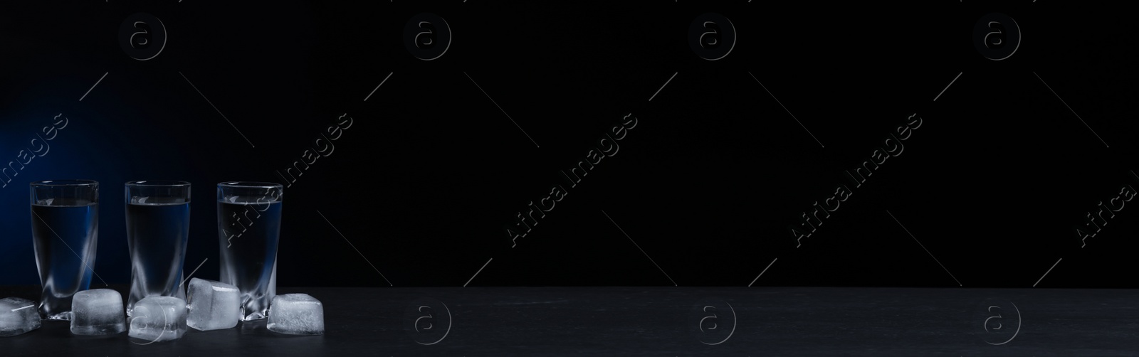 Image of Shot glasses of vodka with ice cubes on black table against dark background, space for text. Banner design