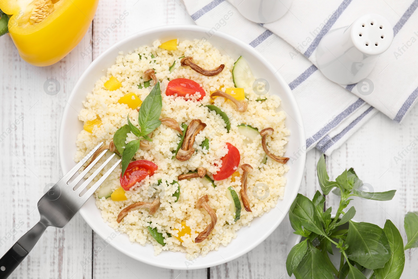 Photo of Bowl of delicious couscous with vegetables and basil served on white wooden table, flat lay