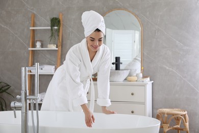 Photo of Beautiful young woman in soft white robe near bathtub at home