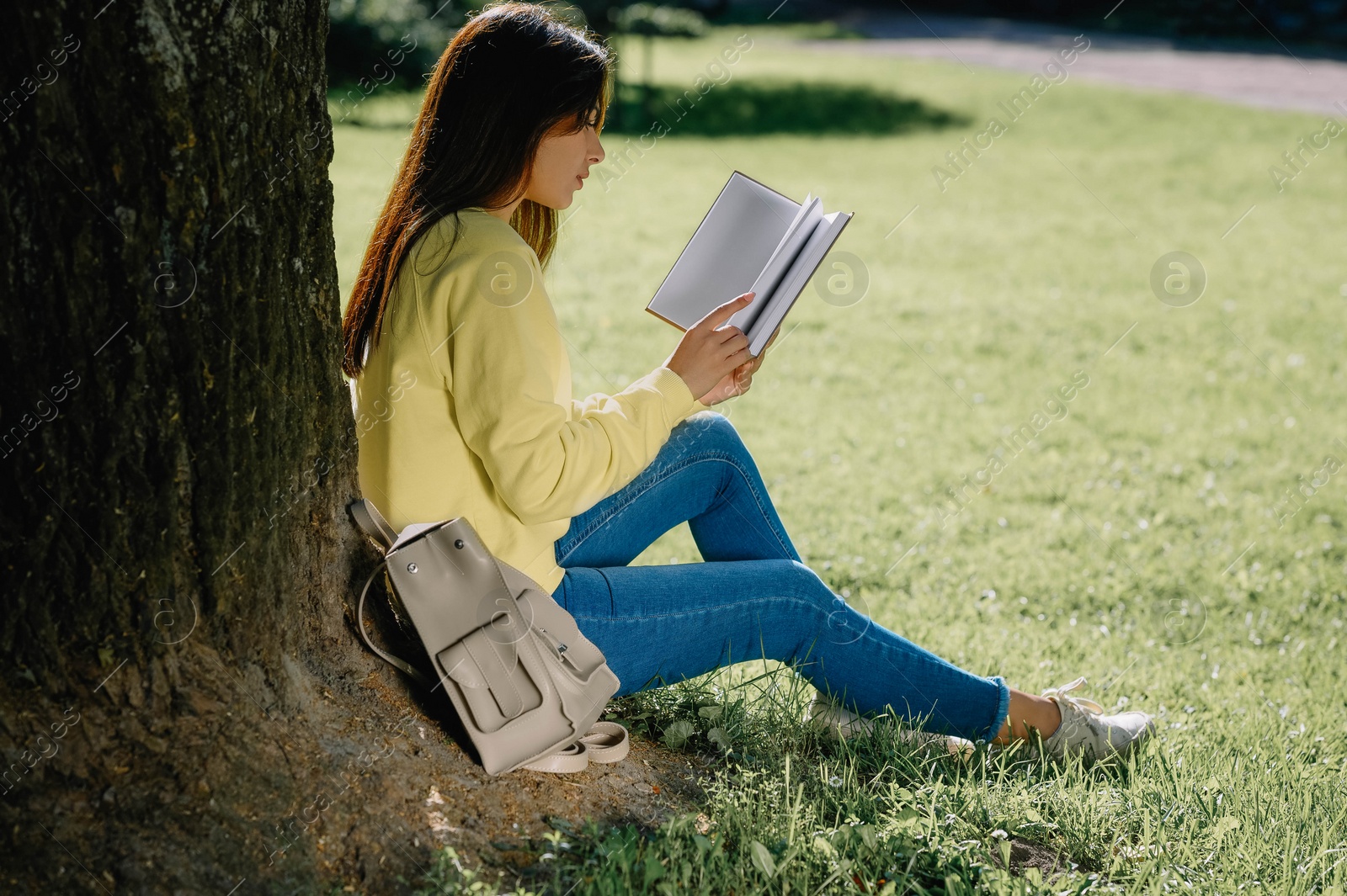 Photo of Young woman reading book near tree in park on sunny day