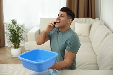 Photo of Emotional man calling roof repair service while collecting leaking water from ceiling in living room