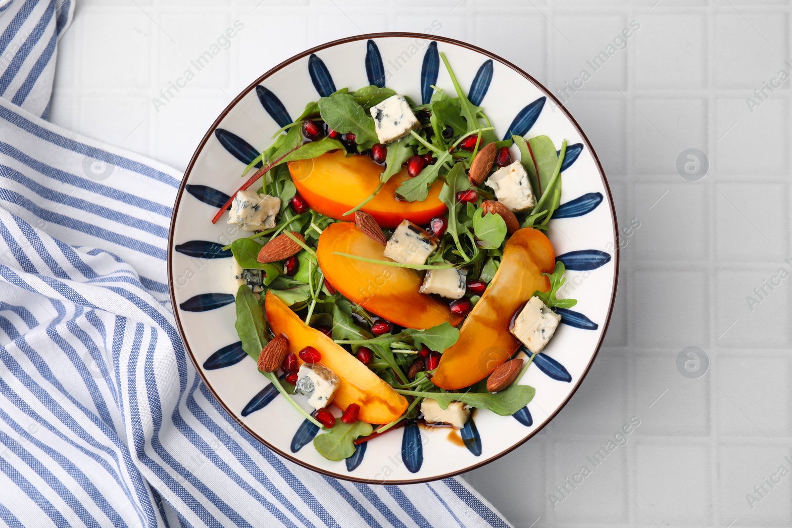 Photo of Tasty salad with persimmon, blue cheese, pomegranate and almonds served on white tiled table, top view