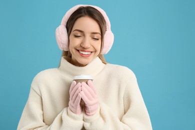 Happy woman with cup of drink wearing warm earmuffs on light blue background