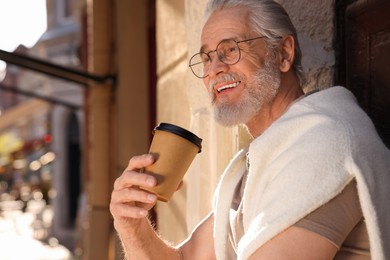 Handsome senior man standing near building with coffee outdoors, space for text