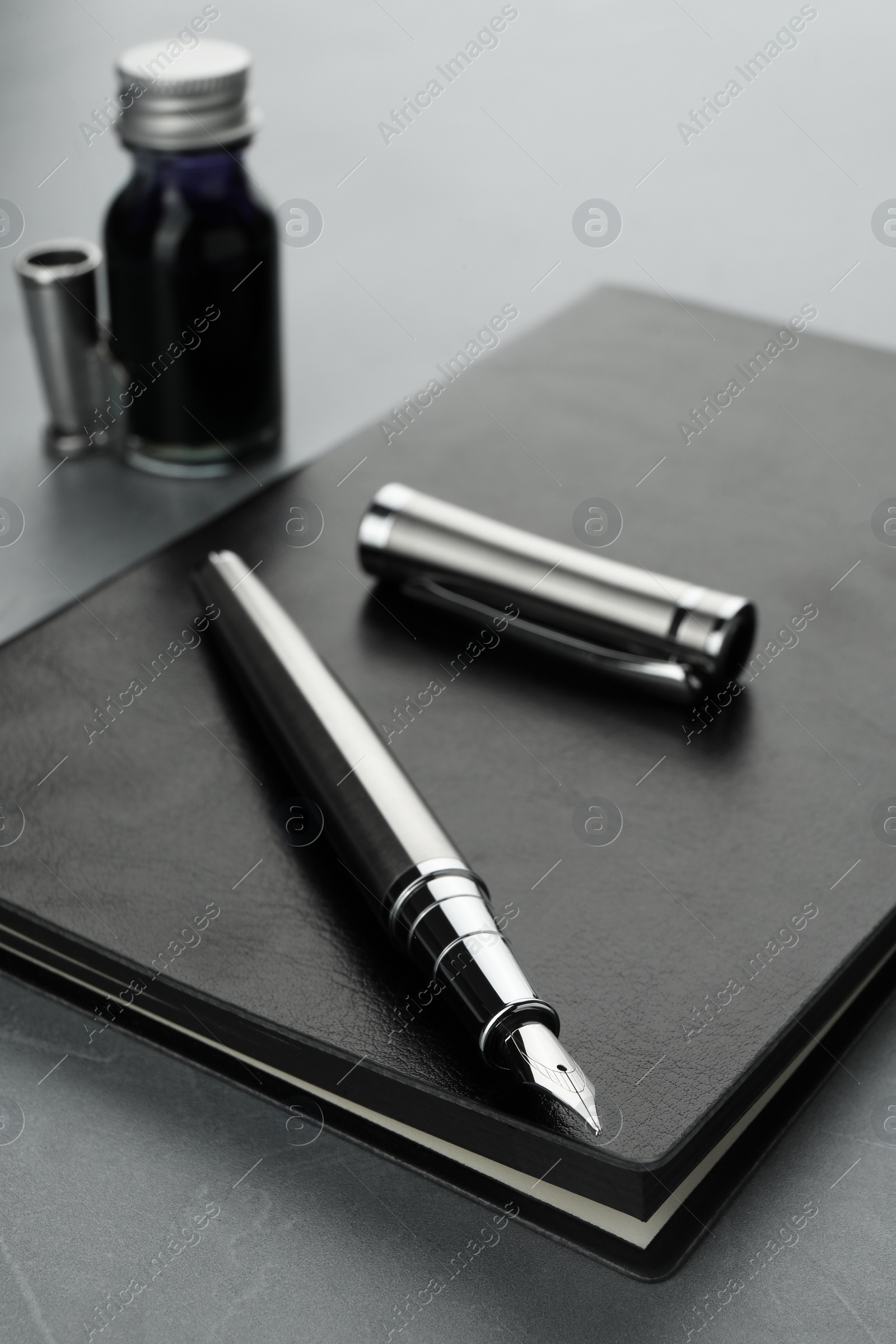 Photo of Stylish fountain pen, bottle of ink, cap and leather notebook on light grey textured table