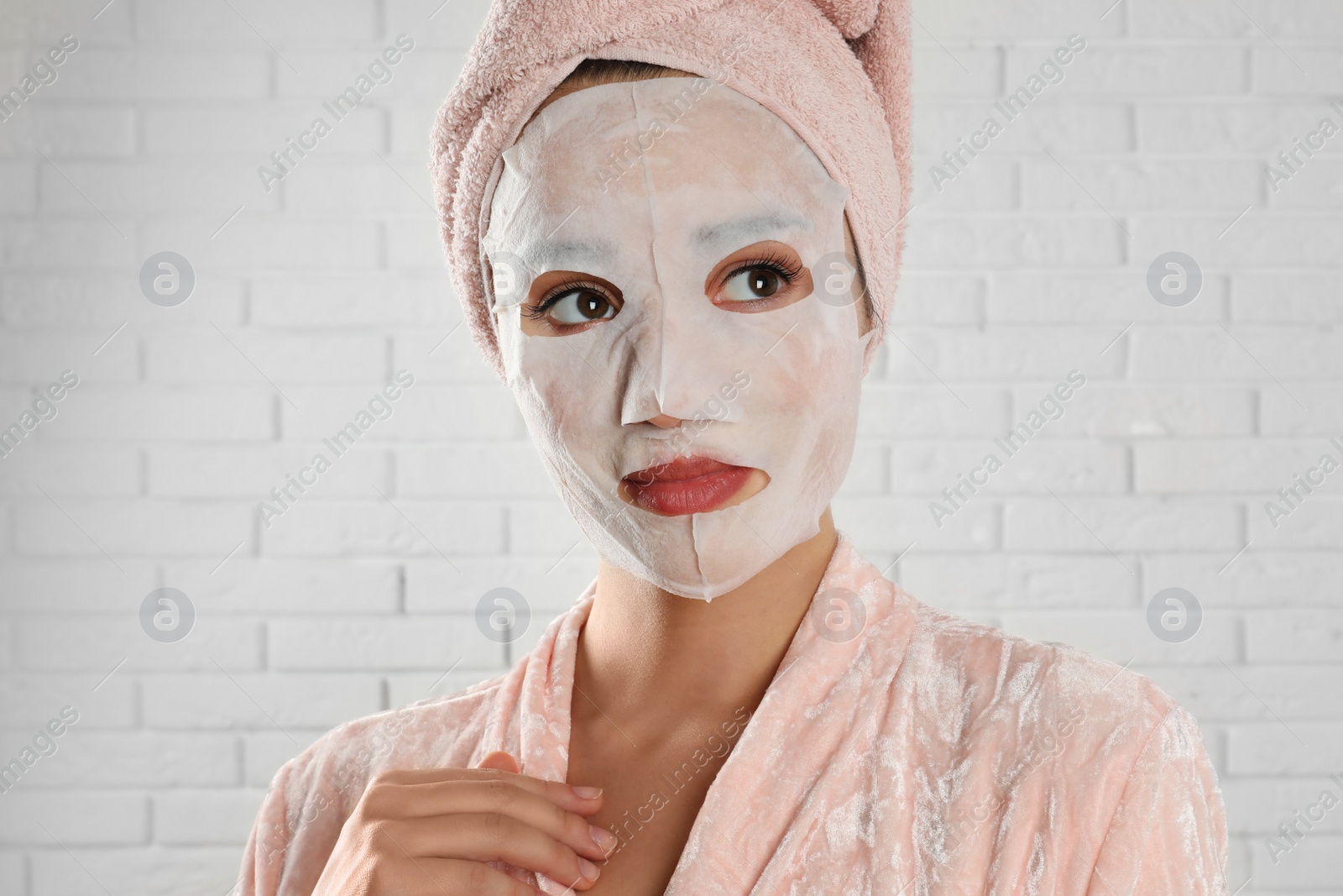 Photo of Young woman in bathrobe with cotton facial mask near white brick wall
