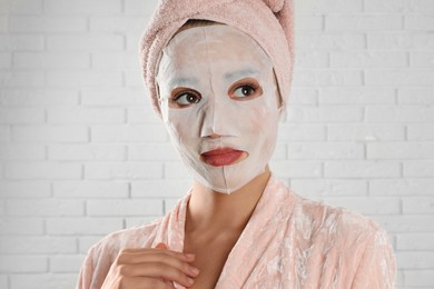 Photo of Young woman in bathrobe with cotton facial mask near white brick wall