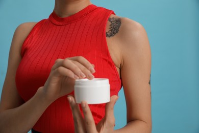 Woman with tattoo holding jar of cream against light blue background, closeup