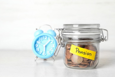 Photo of Coins in glass jar with label "PENSION" on table against light wall. Space for text