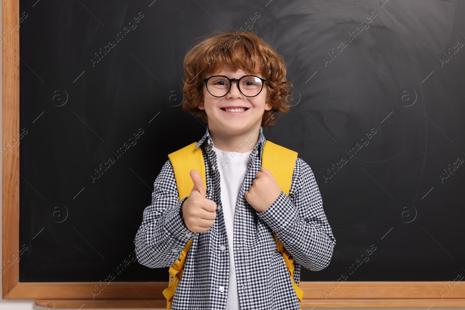 Photo of Happy schoolboy in glasses with backpack showing thumb up gesture near blackboard