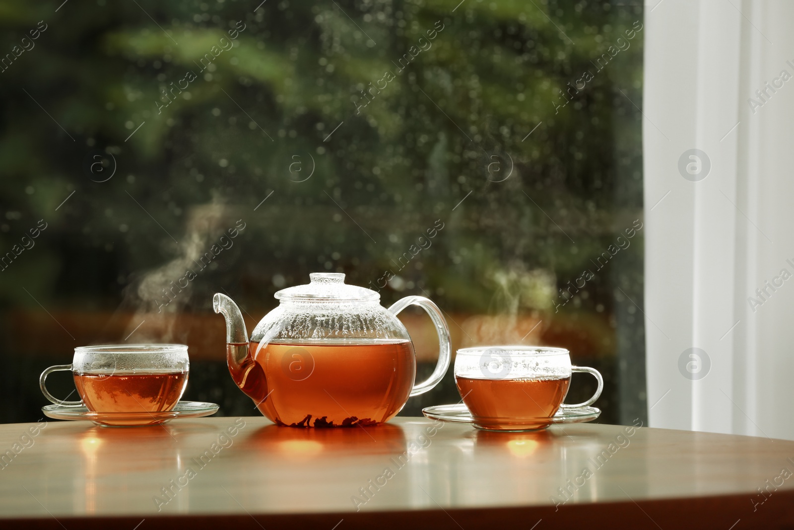 Photo of Teapot and cups of hot tea on wooden table against blurred background, space for text