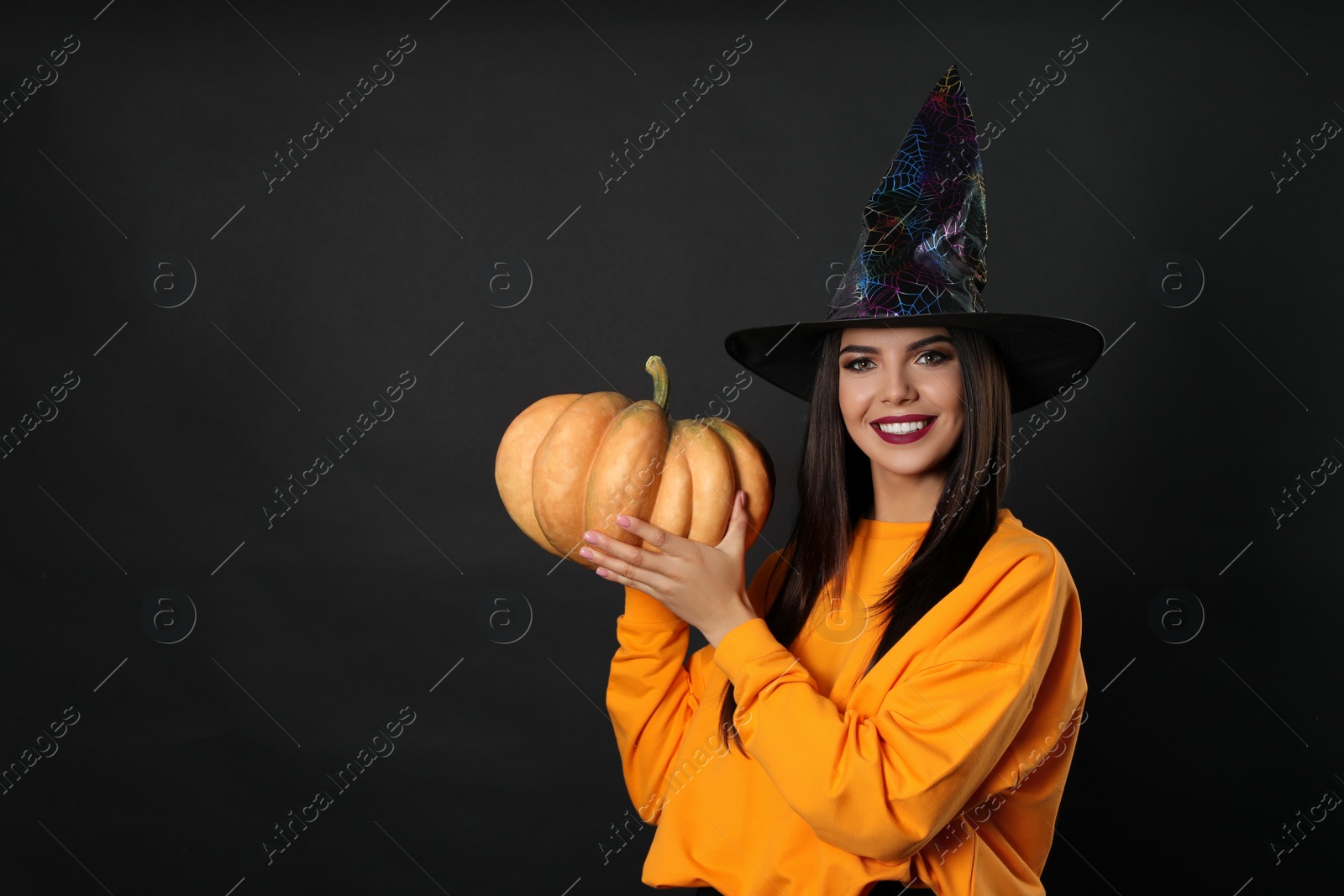 Photo of Beautiful woman wearing witch costume with pumpkin for Halloween party on black background, space for text