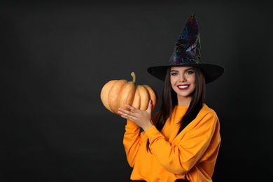 Photo of Beautiful woman wearing witch costume with pumpkin for Halloween party on black background, space for text