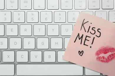 Sticky note with phrase Kiss Me and lipstick mark on keyboard, top view