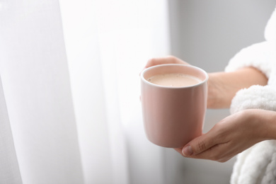 Woman with cup of hot drink near window at home in morning, closeup. Space for text