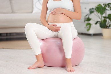 Pregnant woman sitting on fitness ball in room, closeup. Home yoga