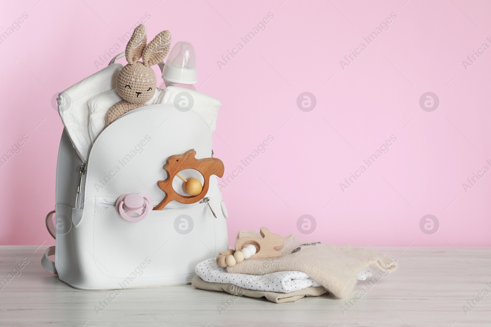 Photo of Mother's backpack with baby's stuff on white wooden table against pink background. Space for text