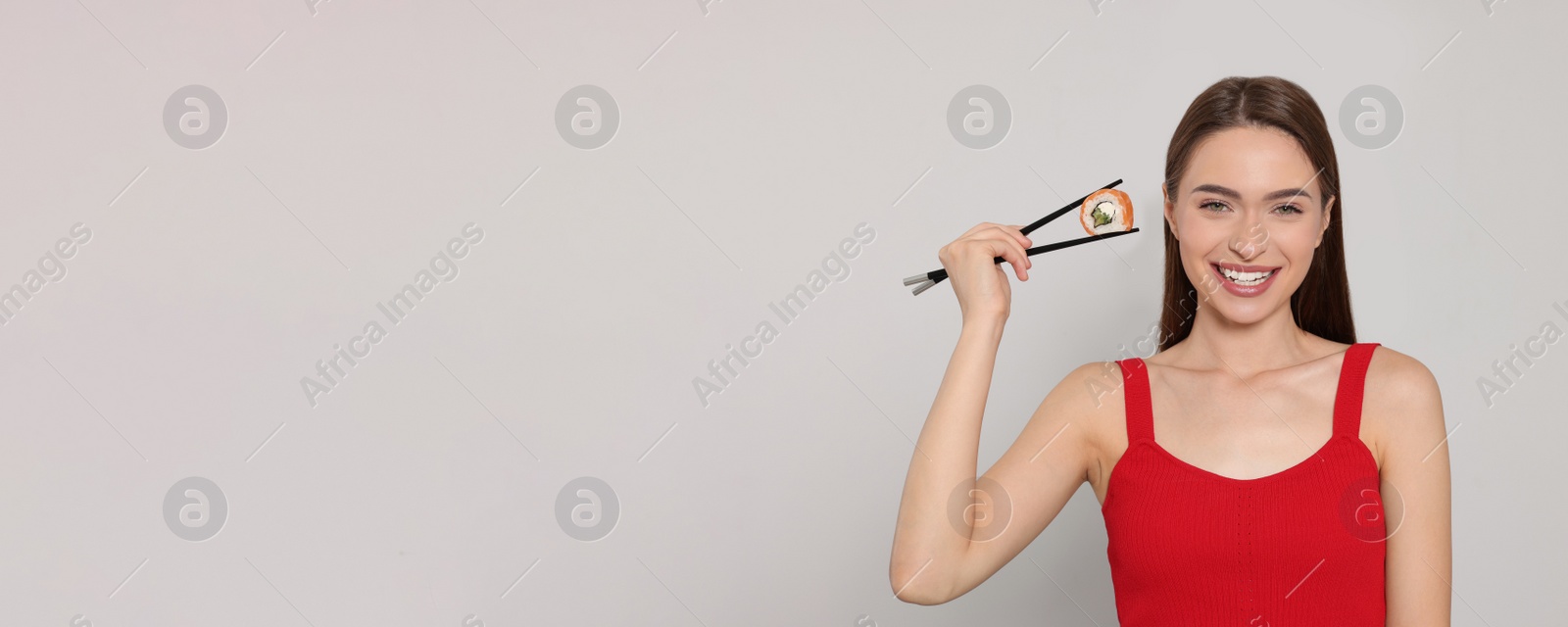 Photo of Happy young woman holding sushi roll with chopsticks on light background. Space for text