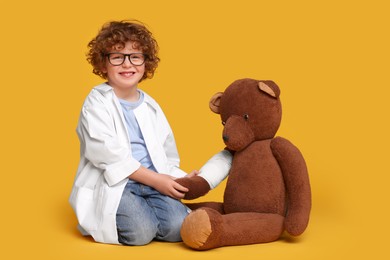 Little boy in medical uniform and toy bear with bandage on yellow background