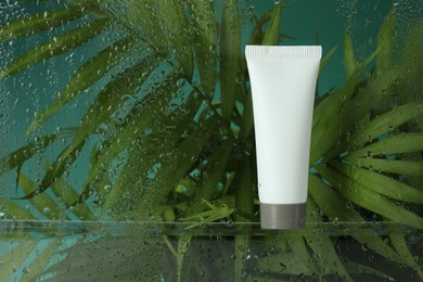 Tube with moisturizing cream and palm leaves on green background, view through wet glass