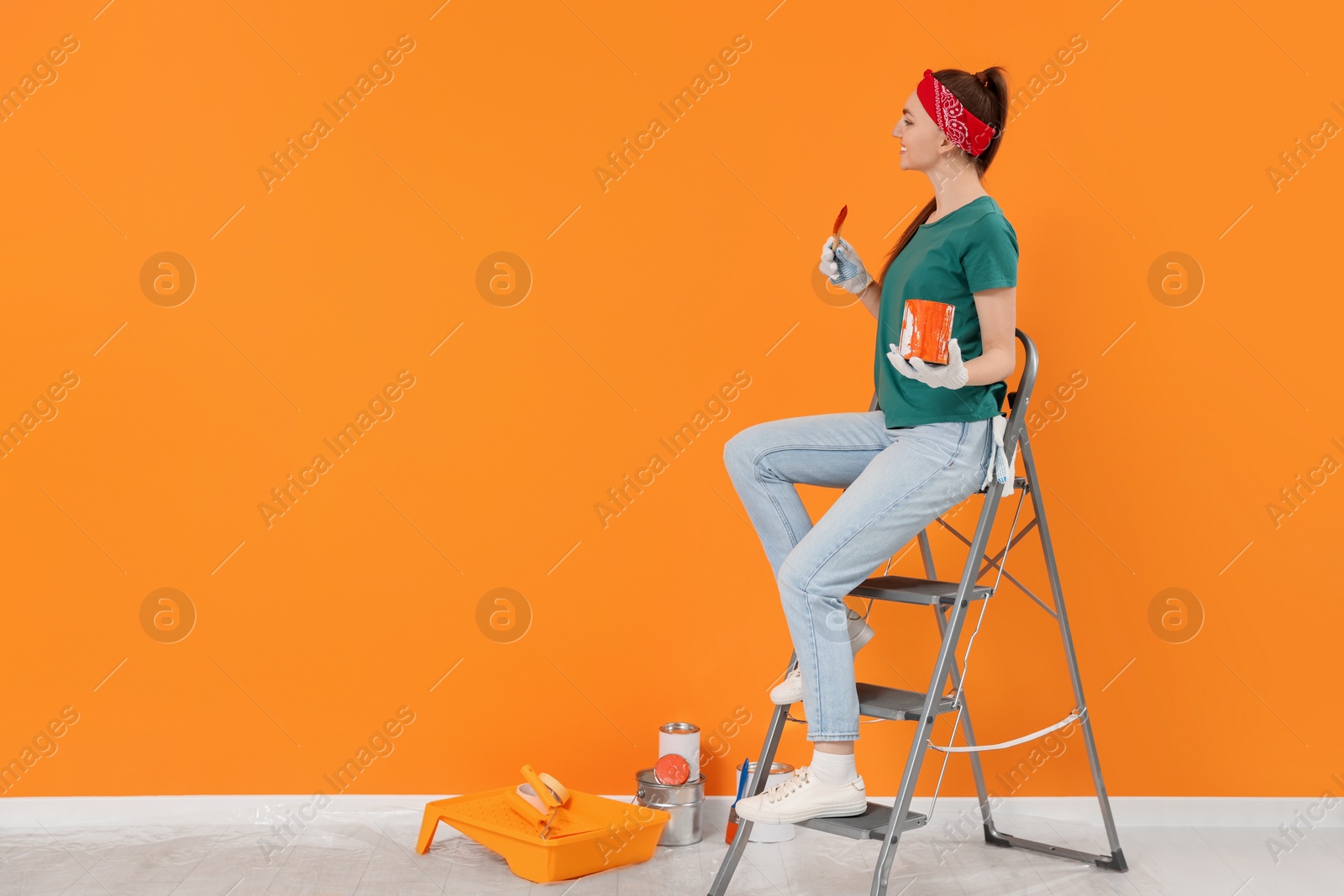 Photo of Designer with painting equipment sitting on folding ladder near freshly painted orange wall indoors, space for text