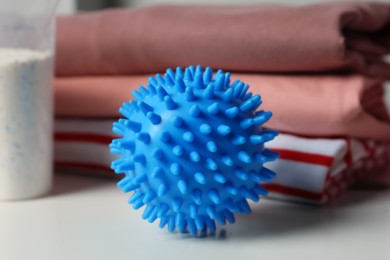 Blue dryer ball, detergent and stacked clean clothes on white table, closeup