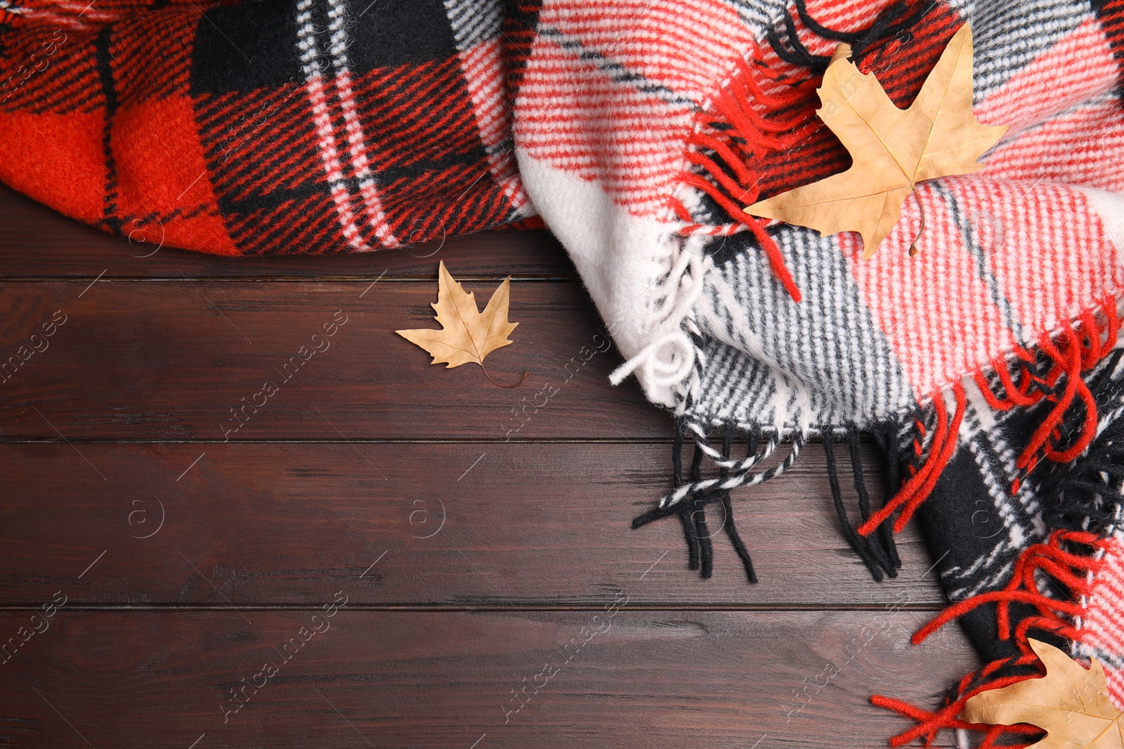 Photo of Red checkered plaid and dry leaves on brown wooden table, flat lay. Space for text