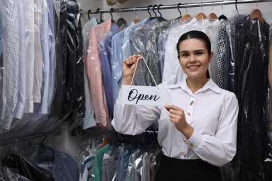 Photo of Dry-cleaning service. Happy worker holding Open sign near rack with clothes indoors, space for text