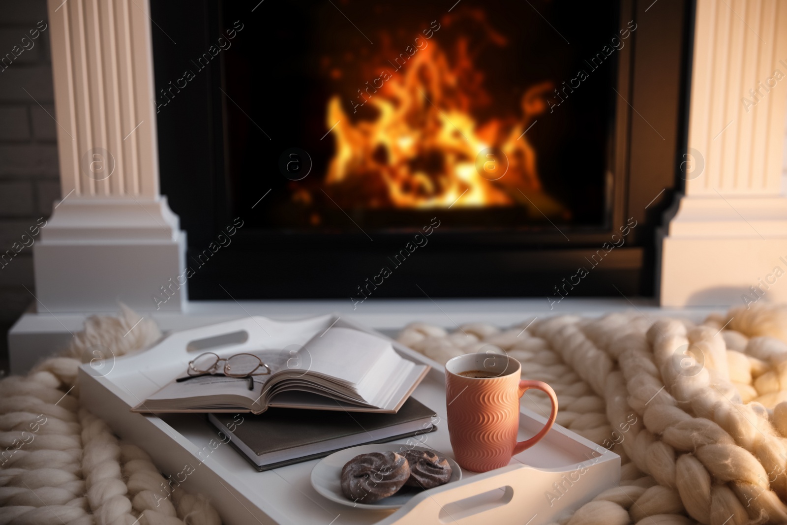 Photo of Cup of coffee, cookies and books on knitted blanket near burning fireplace indoors. Cozy atmosphere