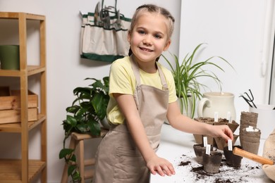 Photo of Little girl inserting cards with names of vegetable seeds into peat pots on window sill indoors