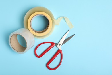 Photo of Rolls of adhesive tape and scissors on light blue background, flat lay. Space for text