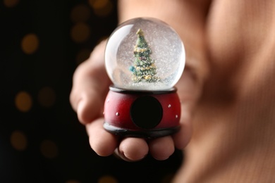 Woman holding snow globe with Christmas tree against festive lights, closeup