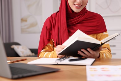 Photo of Muslim woman studying near laptop at wooden table in room, closeup